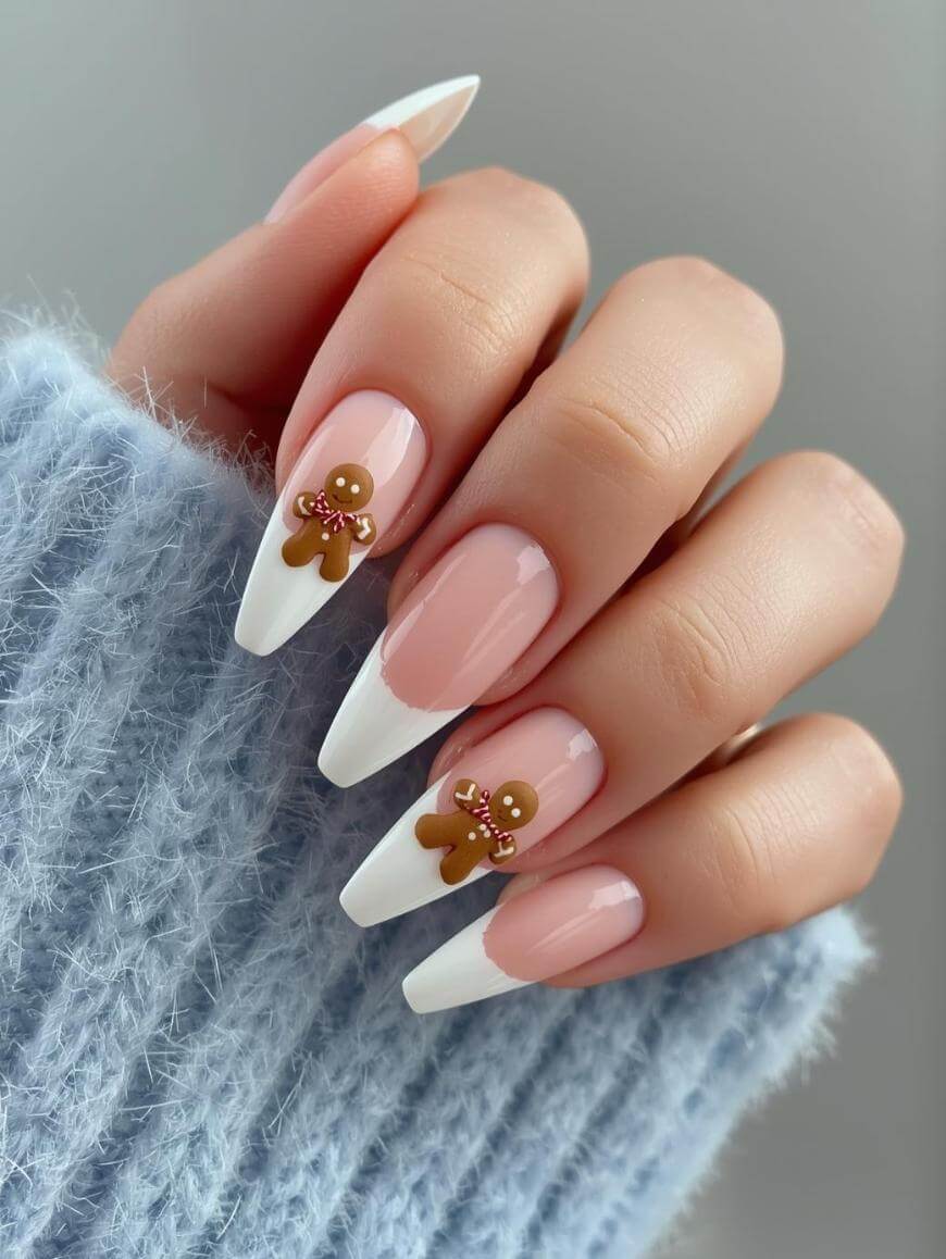 A hand with long, almond-shaped French tip nails features white tips, a pale pink base, and small gingerbread man decorations on two nails. The person is wearing a fuzzy light blue sweater.