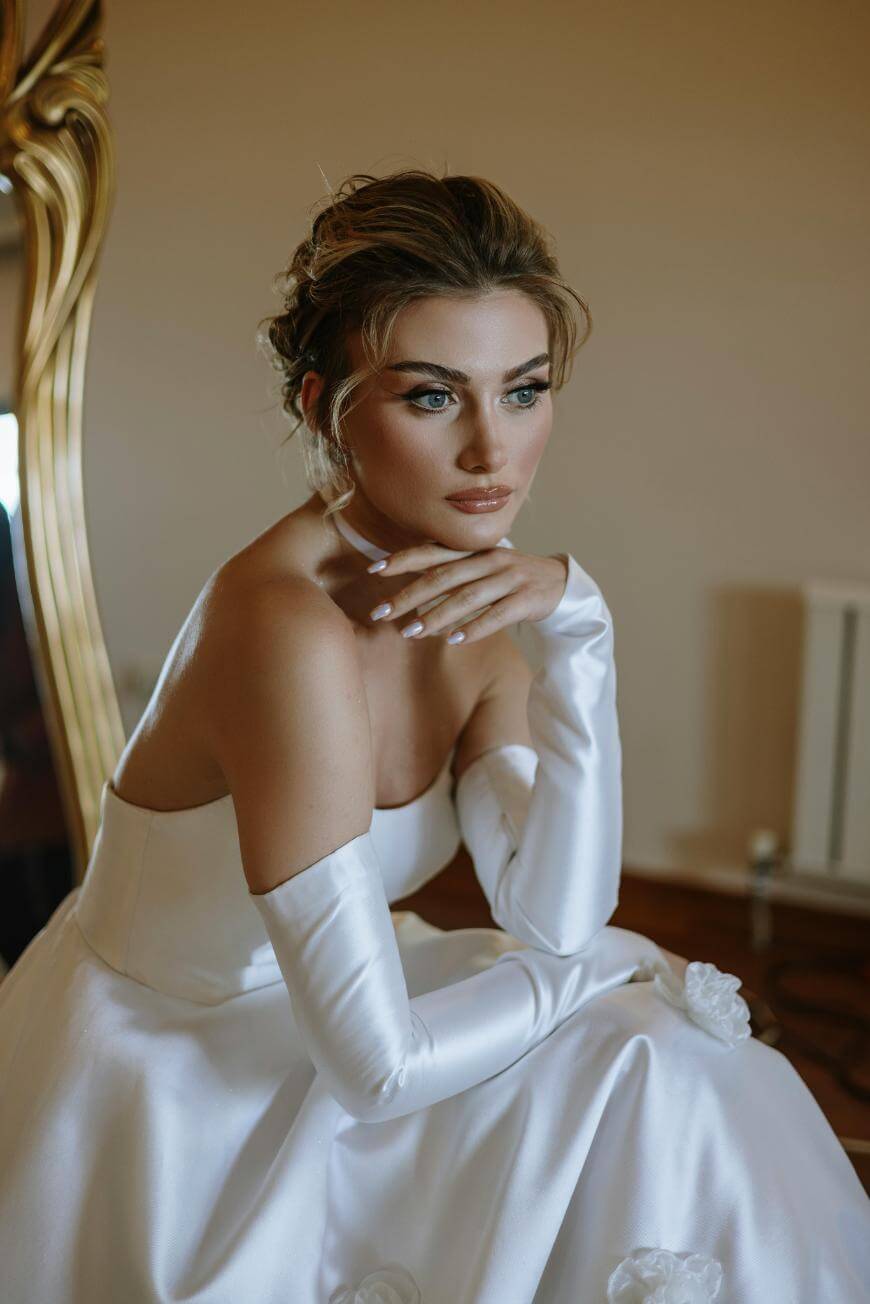 A woman in a white strapless gown and long gloves sits thoughtfully, resting her chin on her hand. She has styled hair and subtle makeup, and is seated indoors near a mirror and soft lighting.