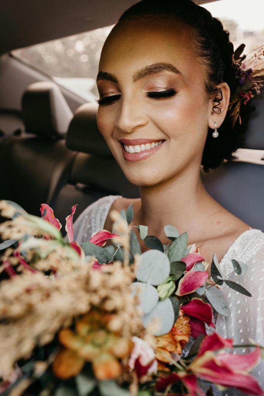 A smiling bride with natural makeup and an elegant updo sits in a car, holding a colorful bouquet of flowers close to her face, eyes closed in a joyful expression.