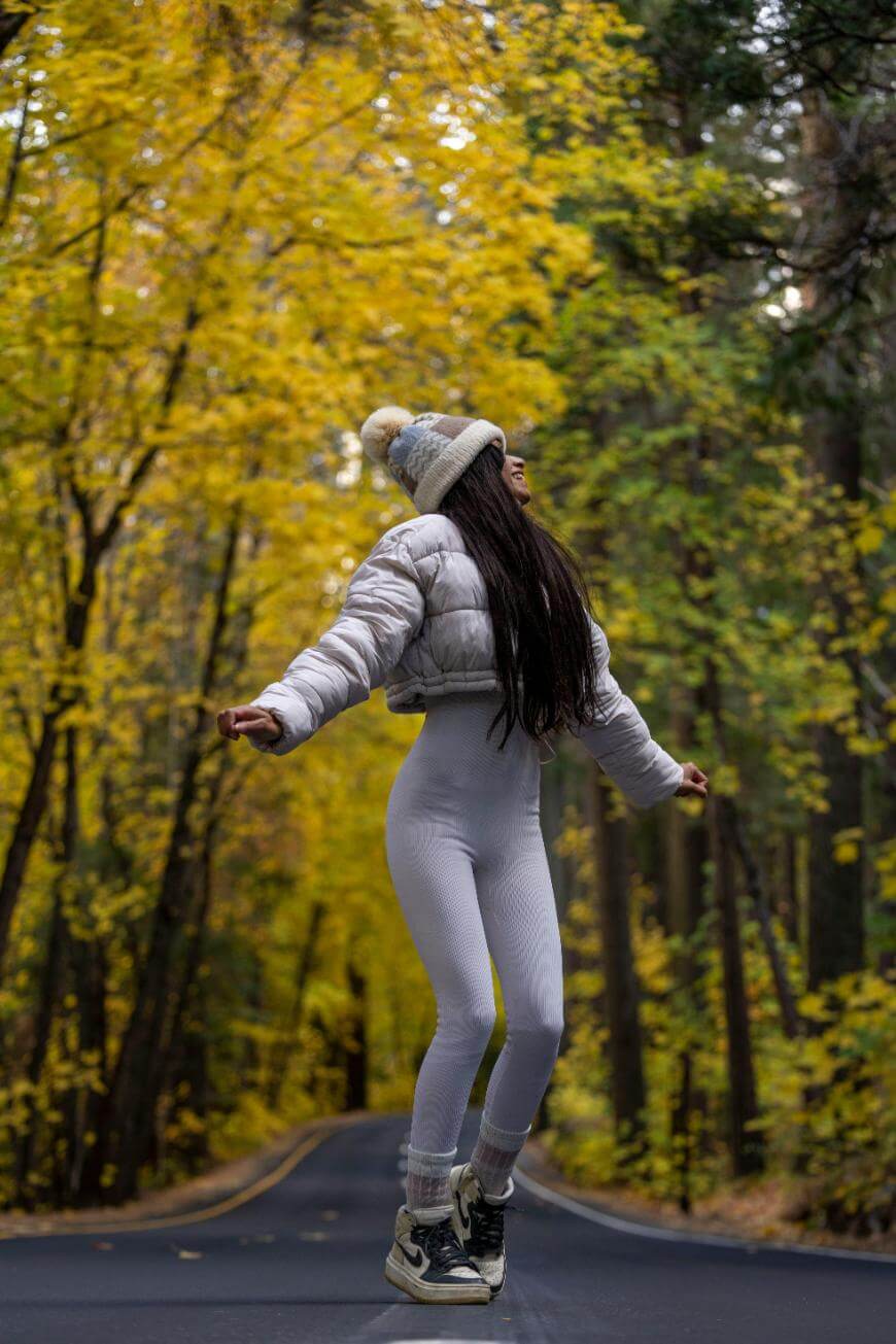 A woman in a white winter outfit and beanie joyfully jumps or skips on a road surrounded by tall trees with vibrant yellow autumn leaves.