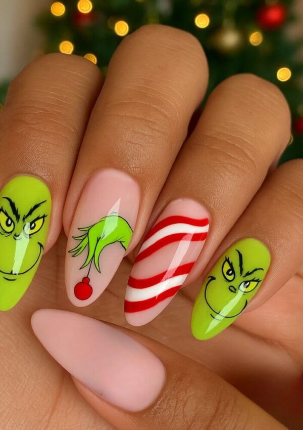 A hand with festive Christmas nail art featuring the Grinch’s face, his green hand holding a red ornament, red and white candy cane stripes, and a plain pale pink nail. A blurred Christmas tree is in the background.
