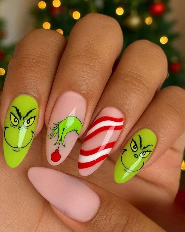 A hand with festive Christmas nail art featuring the Grinch’s face, his green hand holding a red ornament, red and white candy cane stripes, and a plain pale pink nail. A blurred Christmas tree is in the background.