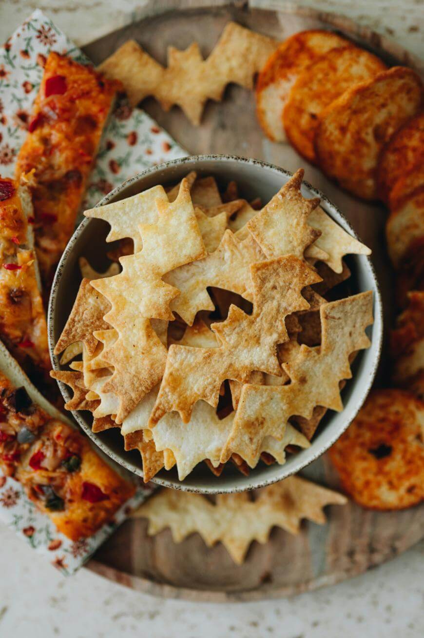 A bowl filled with bat-shaped crackers sits on a wooden board, surrounded by slices of pizza and round, orange-seasoned crackers, creating a festive snack display.