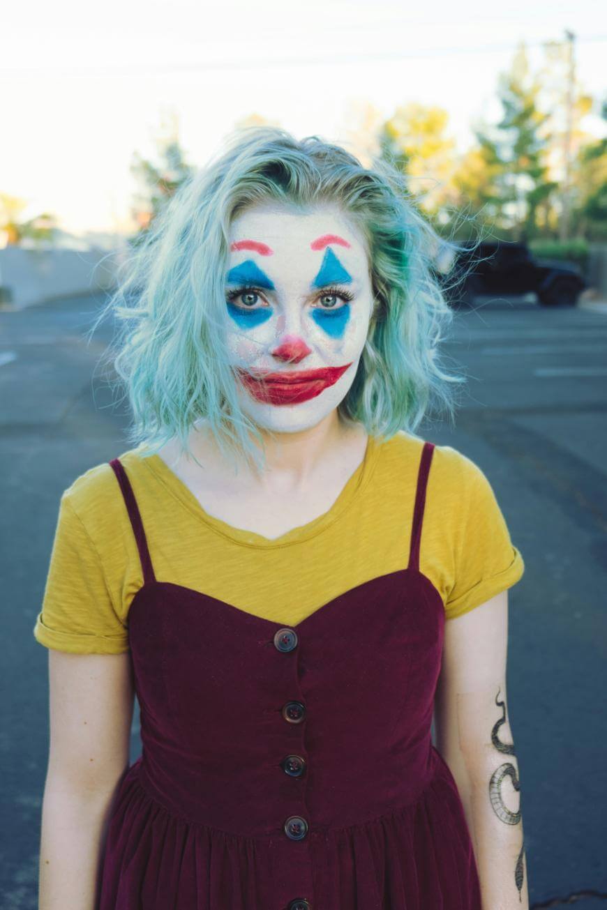 A person with light blue hair and clown makeup stands outdoors, wearing a yellow shirt under a maroon dress. The makeup features blue eyebrows, red lips, and exaggerated eyes. Trees and pavement are visible in the background.