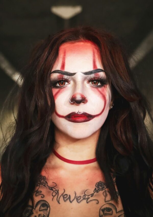 A person with long dark hair wears dramatic clown makeup, featuring red and black lines and a red nose. They have a nose ring, bold lipstick, and visible tattoos on their chest, standing against a blurred background.
