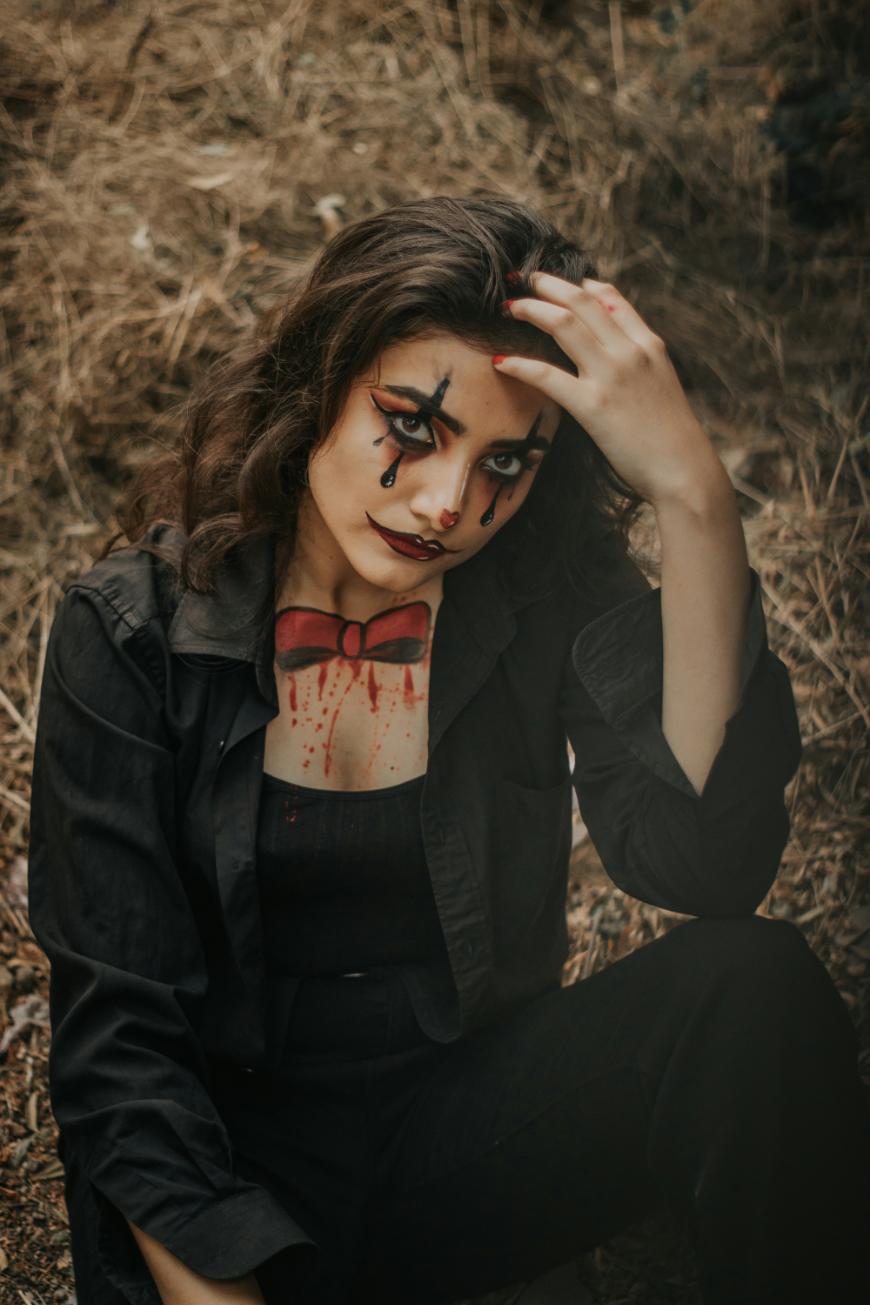 A woman in black clothing sits on the ground outdoors with clown makeup, including painted tears, a red bow tie, and blood-like details on her face and neck, touching her hair with one hand.
