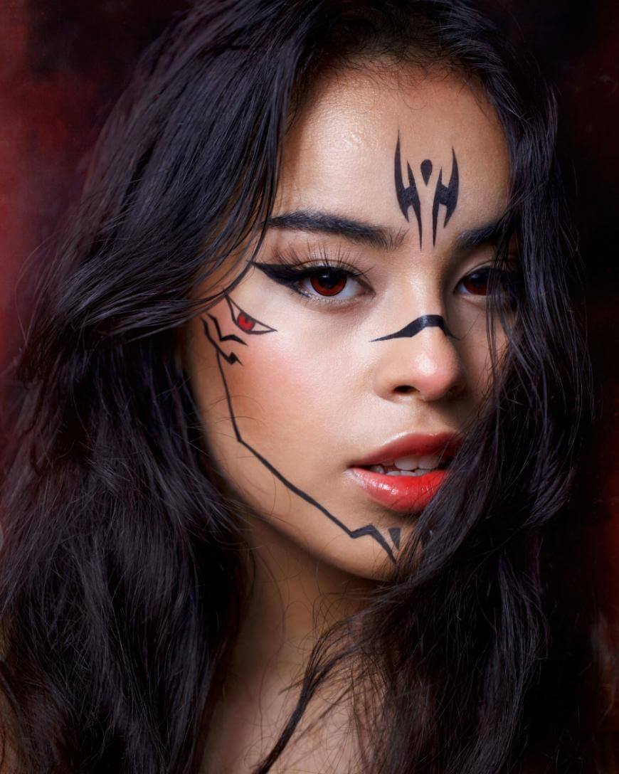A woman with long dark hair faces the camera, her face partly covered by strands of hair. She has dramatic black tribal-style face paint and wears bold makeup with red lipstick and defined eyeliner, set against a dark background.