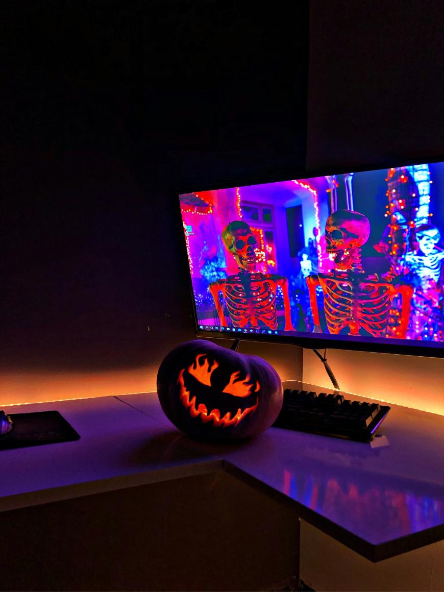A computer desk with a curved monitor displaying two glowing skeletons in a colorful, neon-lit scene. In front of the monitor, a carved pumpkin with a spooky face is illuminated from within, creating a Halloween atmosphere.