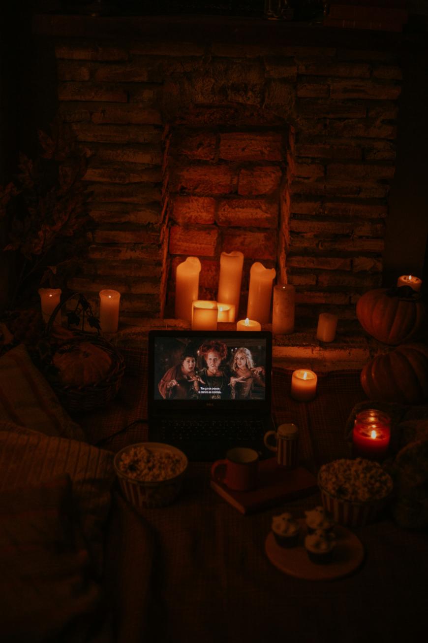 A cozy scene with a laptop playing a movie in front of a fireplace surrounded by glowing candles, pumpkins, popcorn, mugs, and treats, creating a warm, autumn atmosphere.
