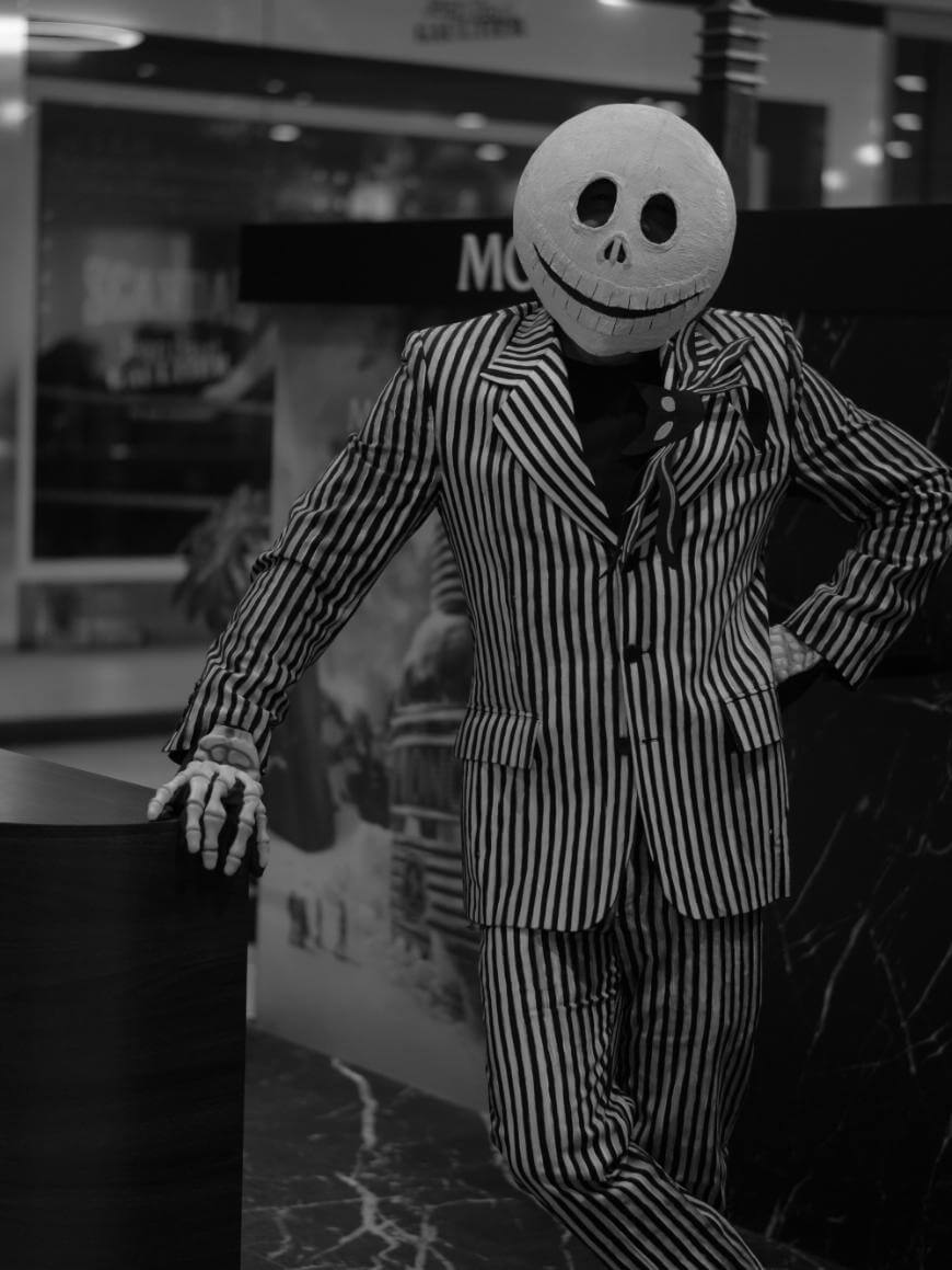 A person wearing a striped suit and a round mask with a wide, toothy grin and large eyes, resembling Jack Skellington from The Nightmare Before Christmas, poses indoors with one hand on a counter. The image is in black and white.
