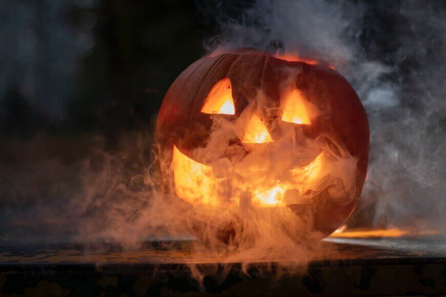 A glowing jack-o’-lantern with a carved smiling face is surrounded by swirling smoke, creating a spooky Halloween atmosphere in dim lighting.