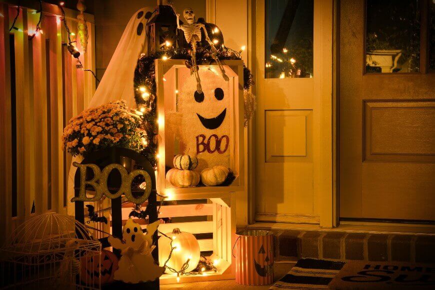 A warmly lit porch decorated for Halloween with string lights, a ghost, pumpkins, a skeleton, flowers, and signs that say BOO. A doormat and festive buckets add to the spooky, welcoming atmosphere by the door.