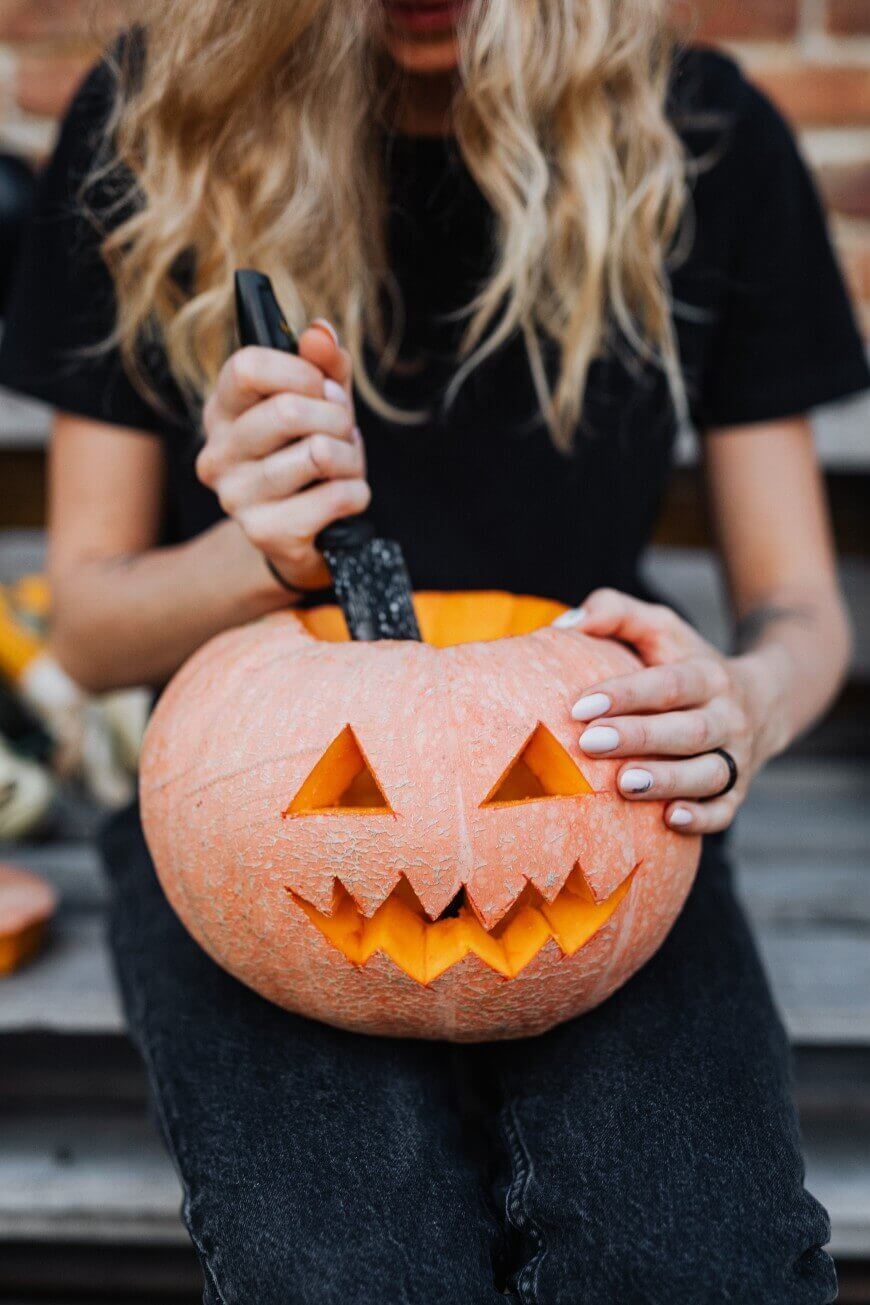 A person with long blonde hair wearing a black shirt carves a jack-o-lantern pumpkin with a knife, sitting on a bench. The pumpkin has triangle eyes and a jagged mouth.