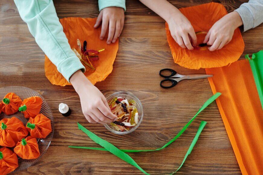Two people making Halloween crafts with orange tissue paper, green paper strips, scissors, and candy on a wooden table. Pumpkin-shaped crafts and a bowl of gummy candy are on the table.
