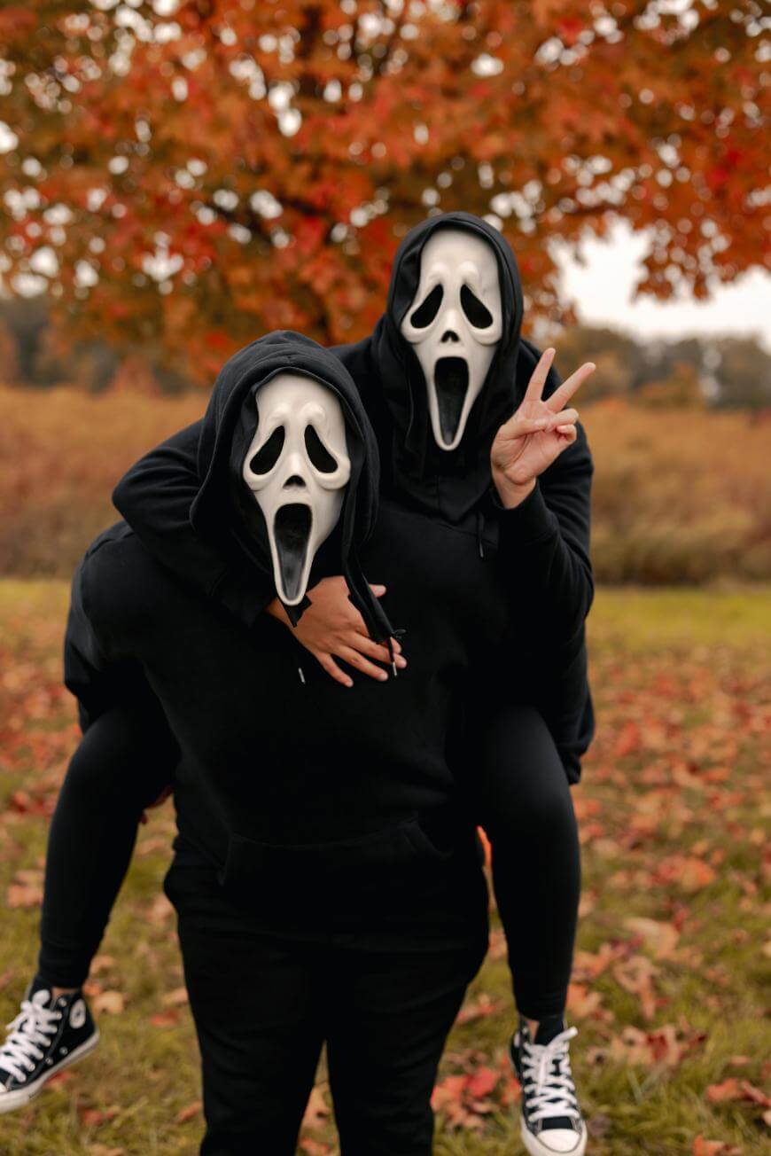 Two people wearing black hoodies and Ghostface masks pose outdoors; one is giving a piggyback ride while the other flashes a peace sign. Autumn leaves and trees with orange foliage are in the background.