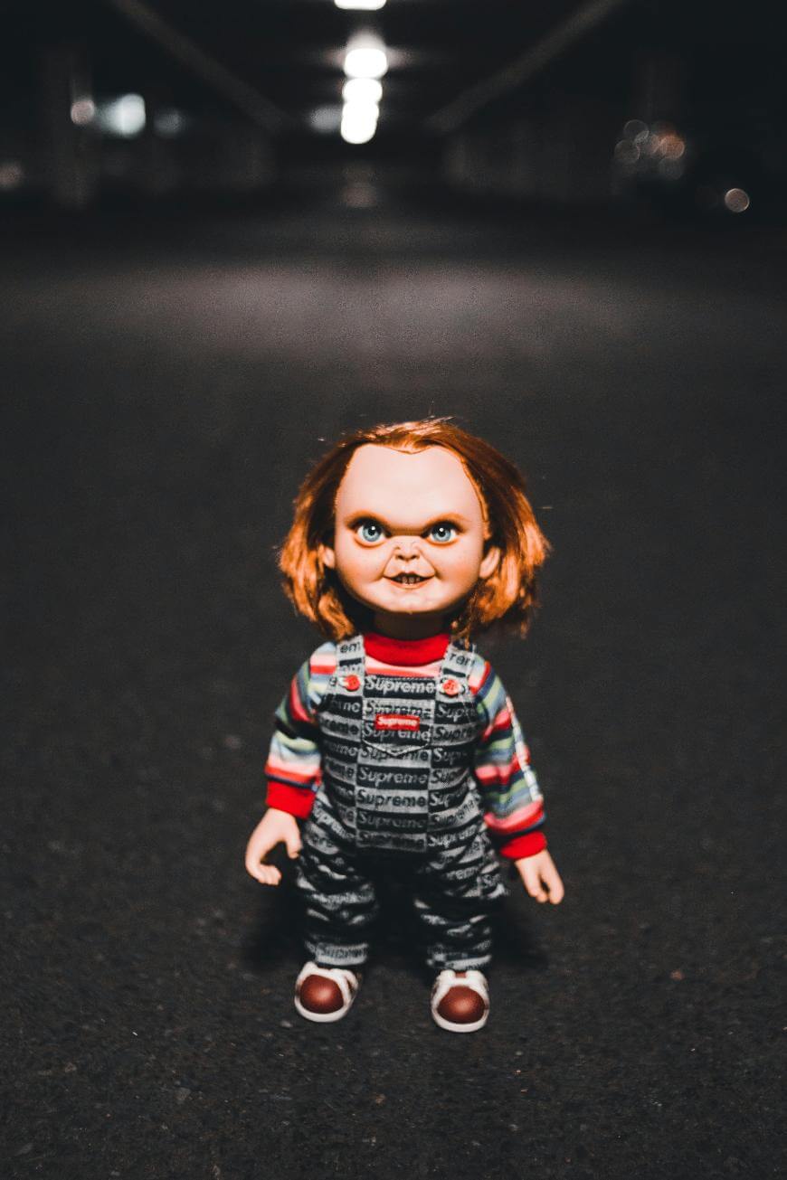 A creepy doll with red hair, wearing a striped shirt and dark overalls, stands alone in the middle of a dimly lit, empty corridor.
