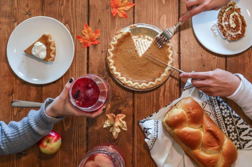 A table set with a pumpkin pie being sliced, a slice of pie with whipped cream, a drink with fruit, an apple, bread, and autumn leaves, with hands reaching for food and drink on a wooden surface.