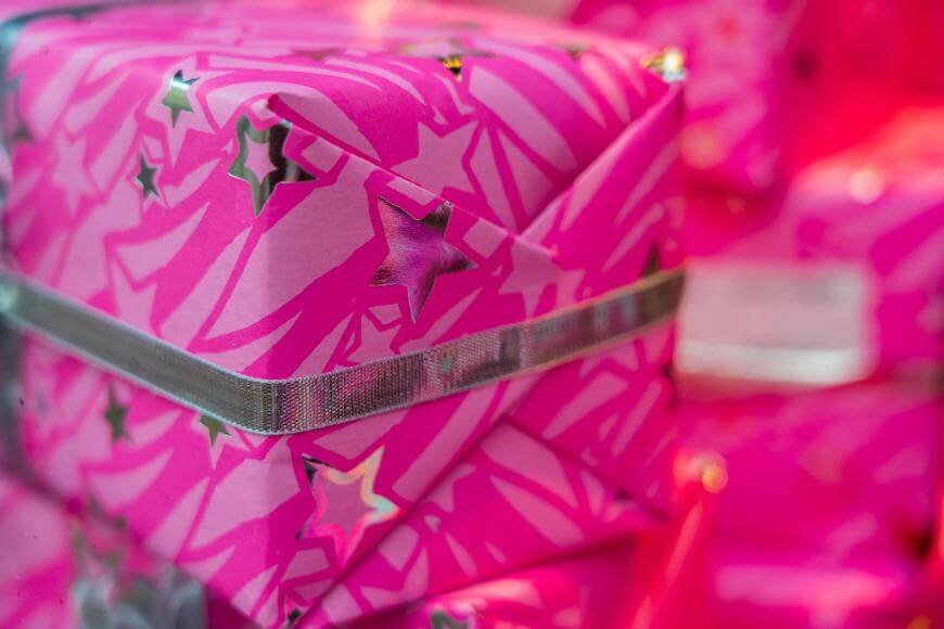 A close-up of a gift wrapped in bright pink paper with shiny silver stars and a silver ribbon, with more similarly wrapped gifts blurred in the background.