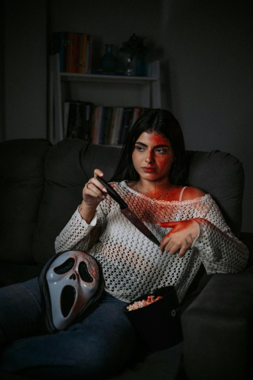 A woman with fake blood on her face sits on a couch, holding a large knife and a bucket of popcorn. A white horror mask rests on her lap, and she is illuminated by a dim, eerie red light.