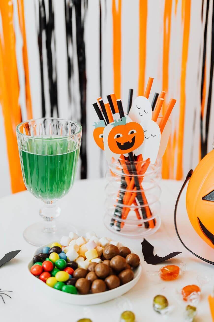 A Halloween-themed table with a green drink in a glass, a bowl of assorted candies, and a jar of black and orange straws decorated with paper pumpkins and ghosts. Orange and black streamers hang in the background.