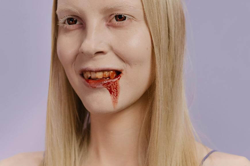 A person with long blonde hair and pale skin smiles, revealing fake vampire fangs and blood dripping from their mouth. Their eyebrows are very light, and they have a mysterious, eerie look. The background is light purple.