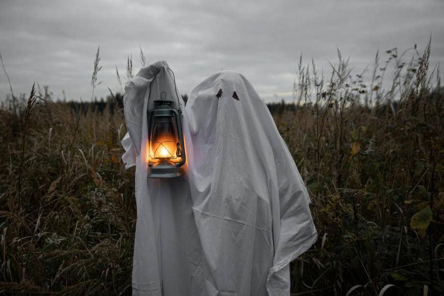A person dressed as a ghost with a white sheet over their body holds a lit lantern, standing in a field of tall grass under a cloudy, gray sky.