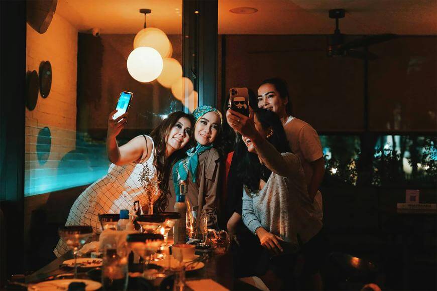 Four women are gathered around a table at a dimly lit restaurant, smiling and posing for group selfies with their phones. Warm light and reflections from the window create a cozy, cheerful atmosphere.