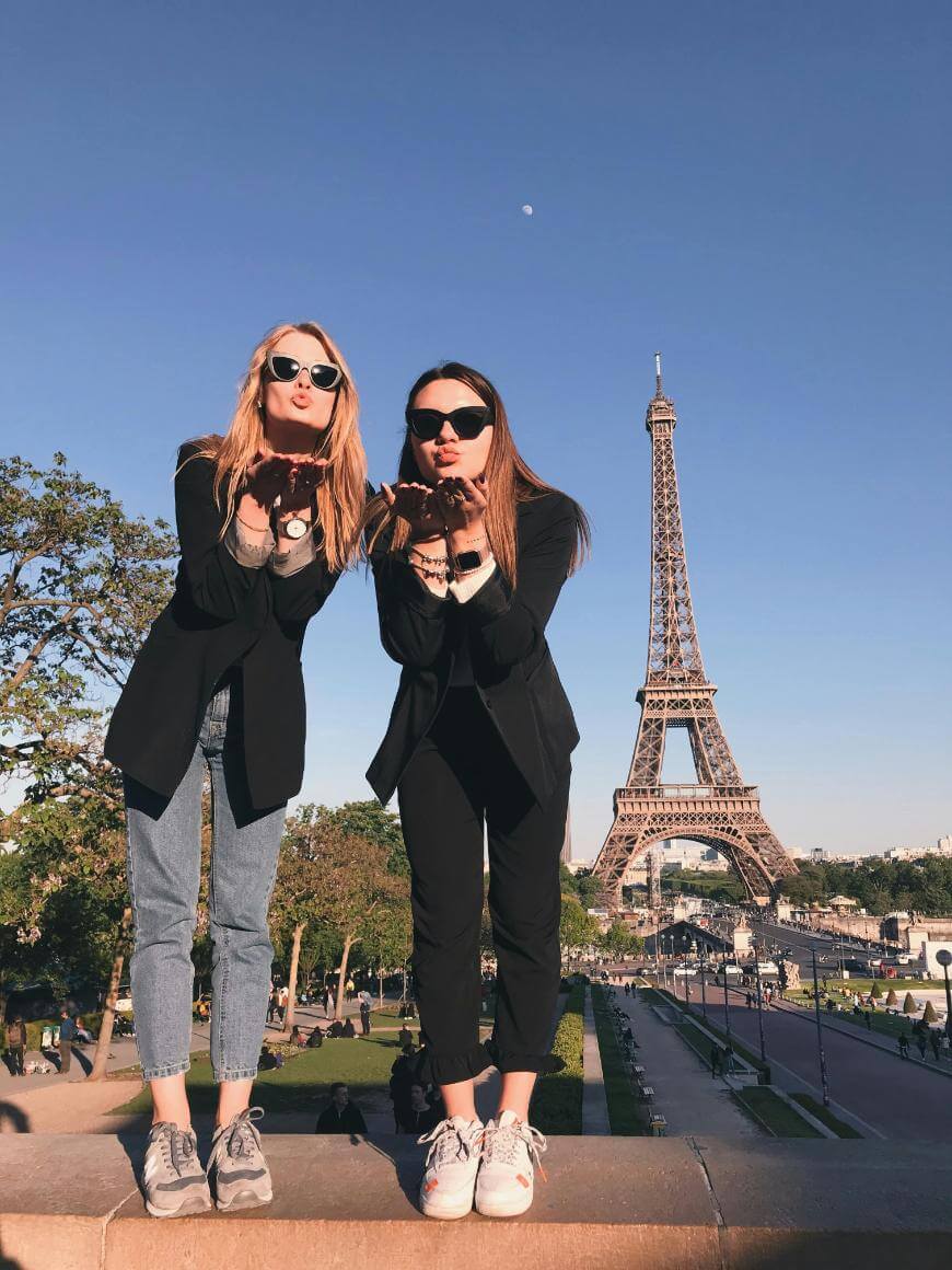 Two women wearing sunglasses and casual clothes stand on a ledge, blowing kisses toward the camera. The Eiffel Tower and a clear blue sky are visible in the background.