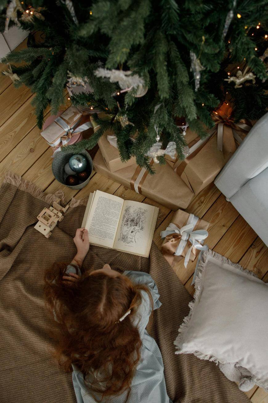 A person with long, wavy hair reads a book while lying on a blanket next to a decorated Christmas tree with wrapped gifts underneath. Ornaments, a wooden toy, and a pillow are nearby on a wooden floor.