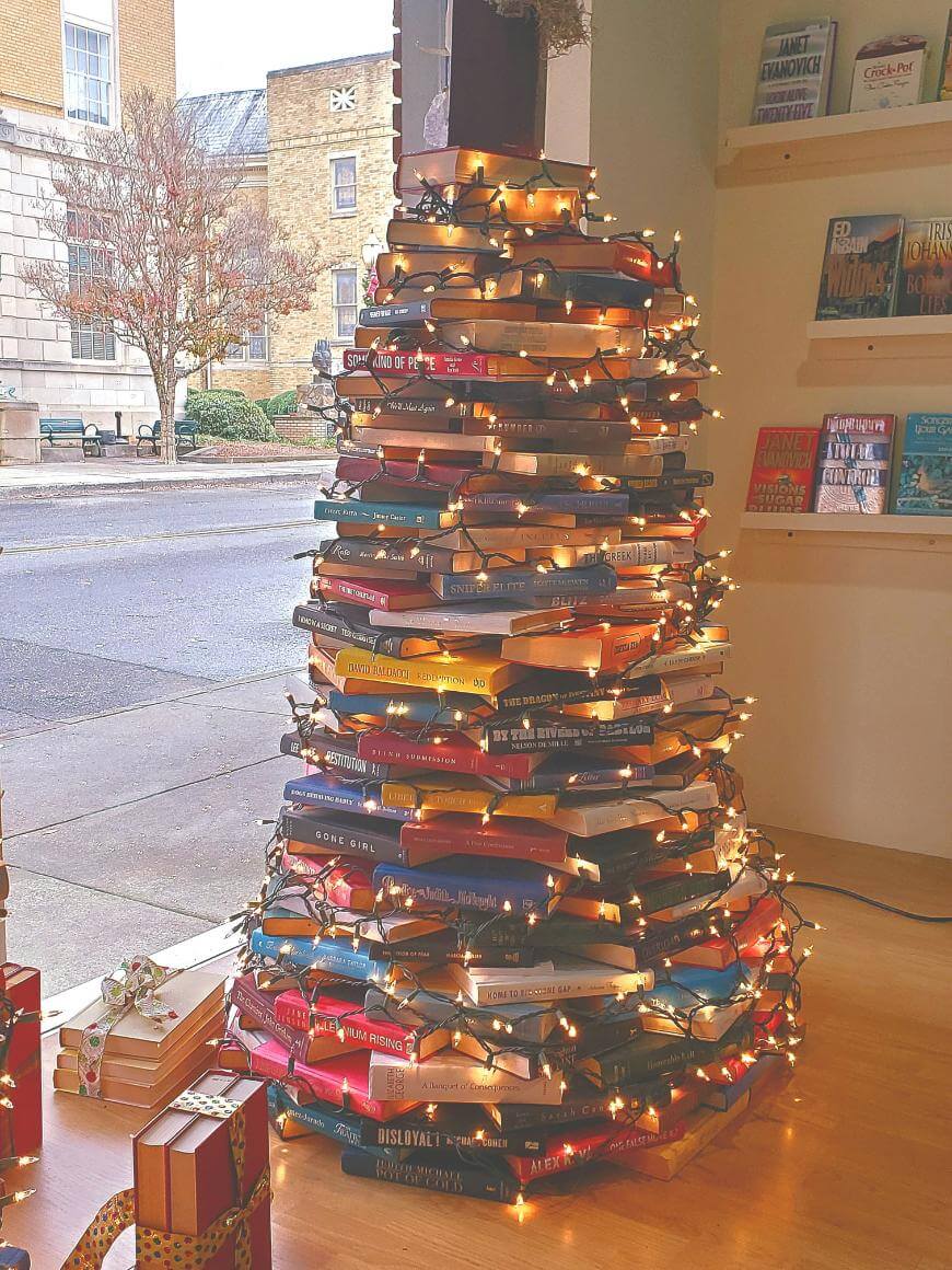 A Christmas tree made from stacked books is decorated with string lights and placed near a window. Wrapped gifts sit beside it, and books are displayed on nearby shelves with a street view visible outside.