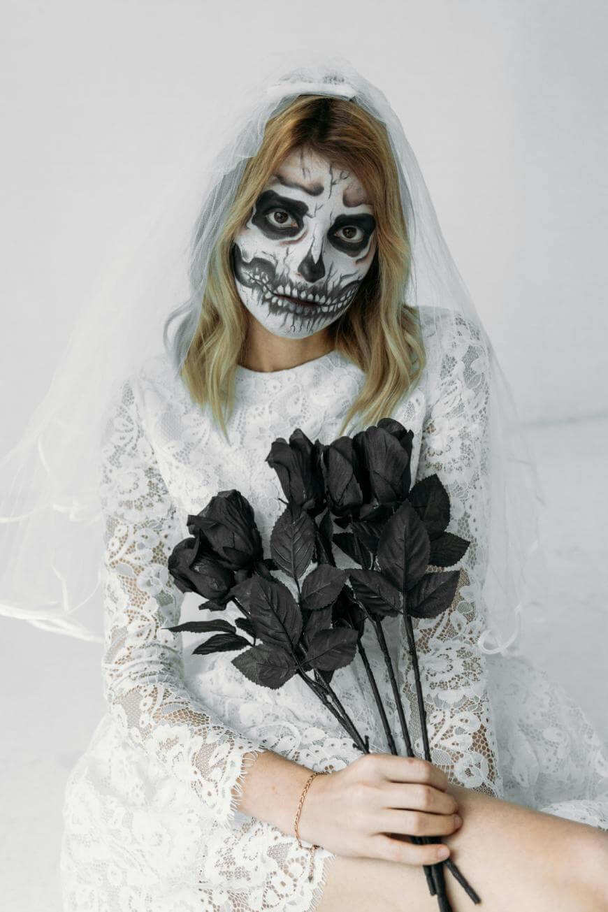 A person in a lace dress and veil, with dramatic skull face paint, holds a bouquet of black roses against a plain white background.