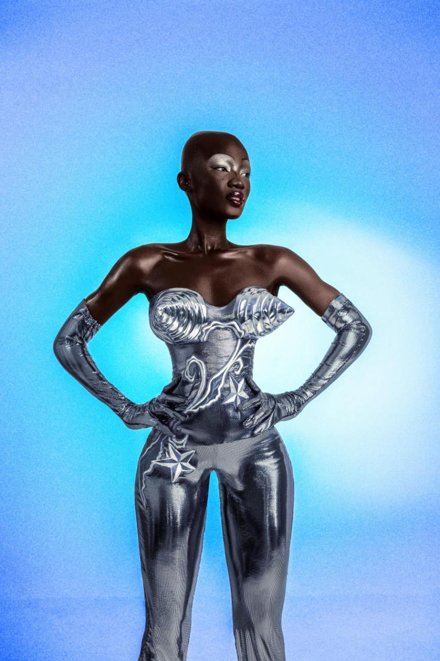 A woman in a futuristic, metallic silver outfit with sculpted shell and star details poses confidently against a bright blue gradient background. She wears matching long gloves and pants, and stands with her hands on her hips.