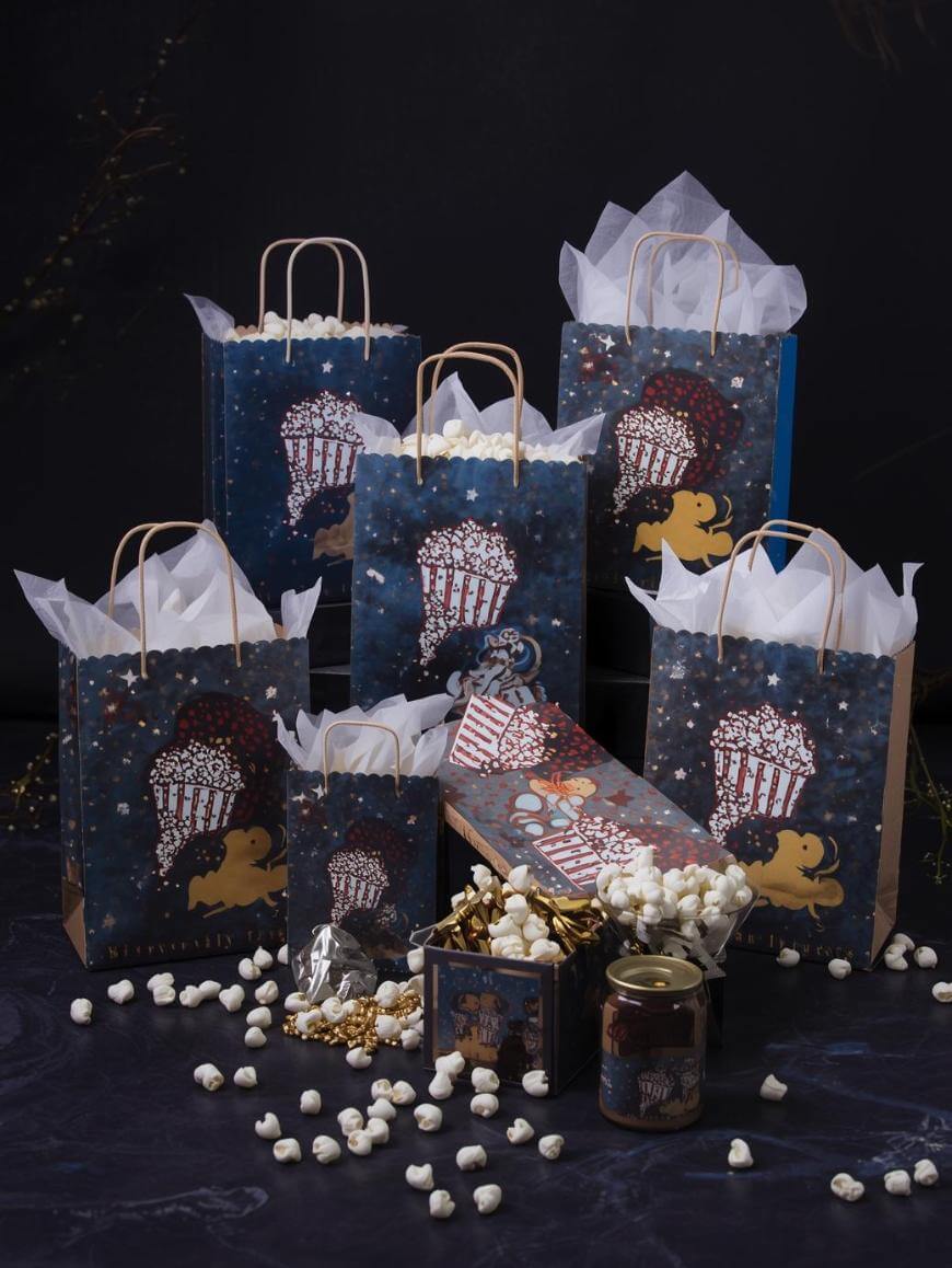 Several gift bags with popcorn-themed designs, tissue paper, and scattered popcorn kernels are displayed on a dark surface, along with matching boxes, a jar, and gold ribbon, creating a festive arrangement.