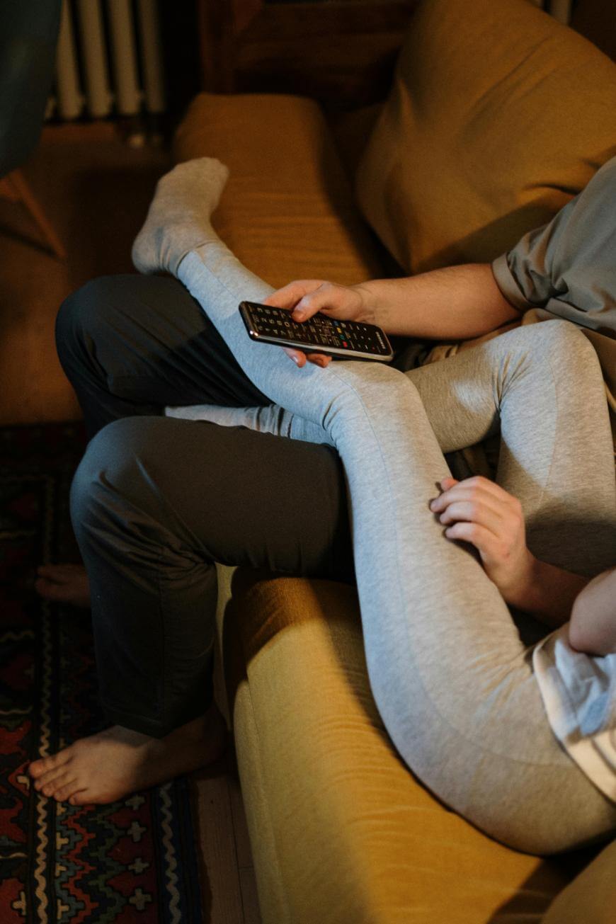Two people relax on a yellow couch, one person’s head resting on the others lap. They wear comfortable clothes and one is holding a TV remote. Their legs are intertwined and the scene feels cozy and casual.