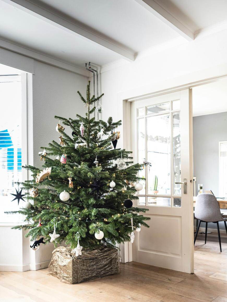 A decorated Christmas tree stands in a bright living room near glass doors, adorned with white and gold ornaments, lights, and a tree skirt at the base. Sunlight streams in through nearby windows.