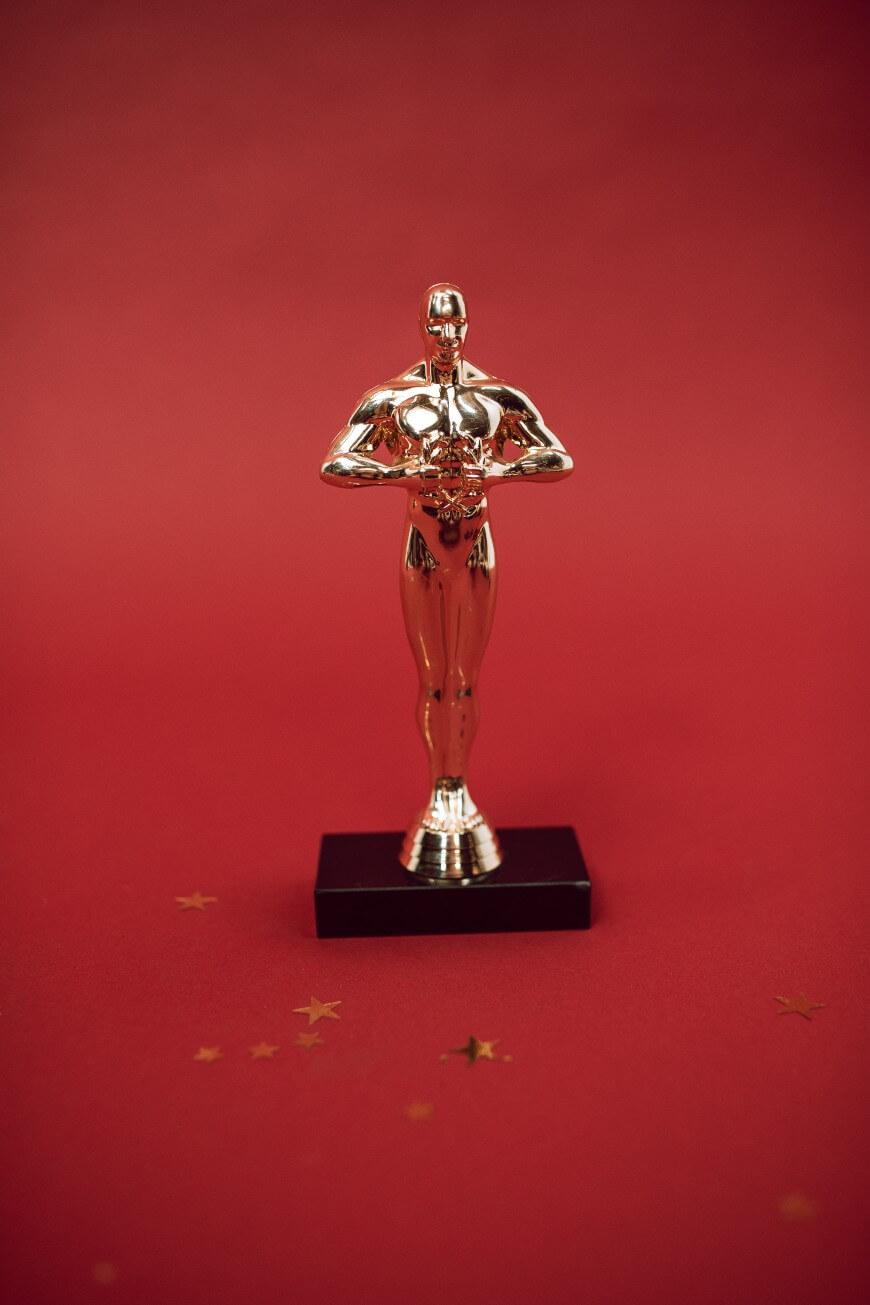 A gold trophy shaped like a human figure stands on a black base against a red background with a few scattered gold star confetti.
