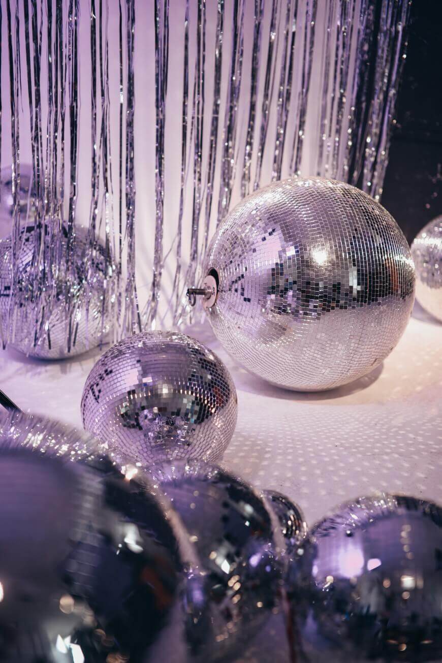 Several large and small disco balls reflect light, surrounded by shiny silver tinsel curtains, creating a festive and sparkling atmosphere.