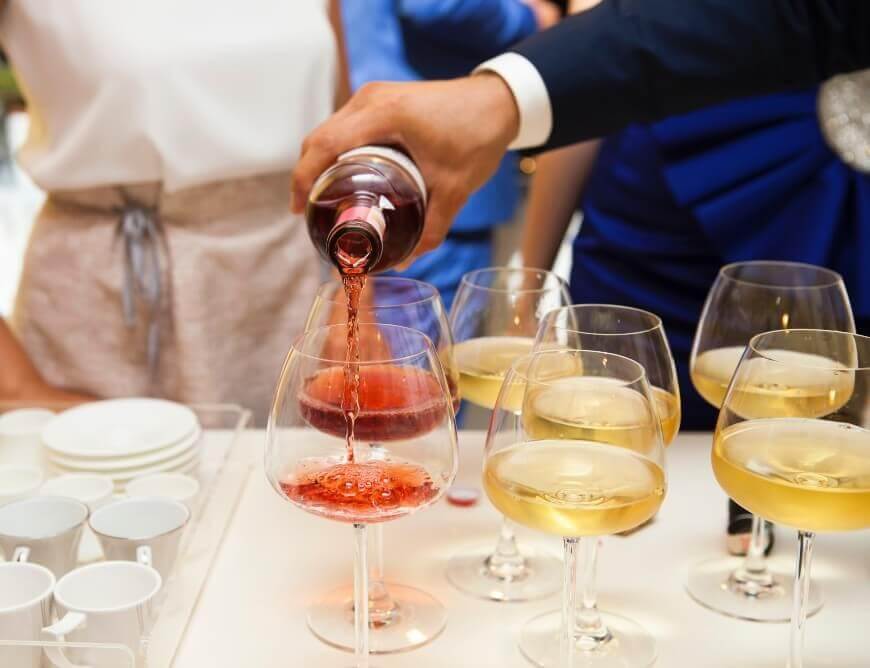 A person pours red wine into a glass among several glasses of white wine on a table, with people standing nearby and stacked white cups visible in the background.