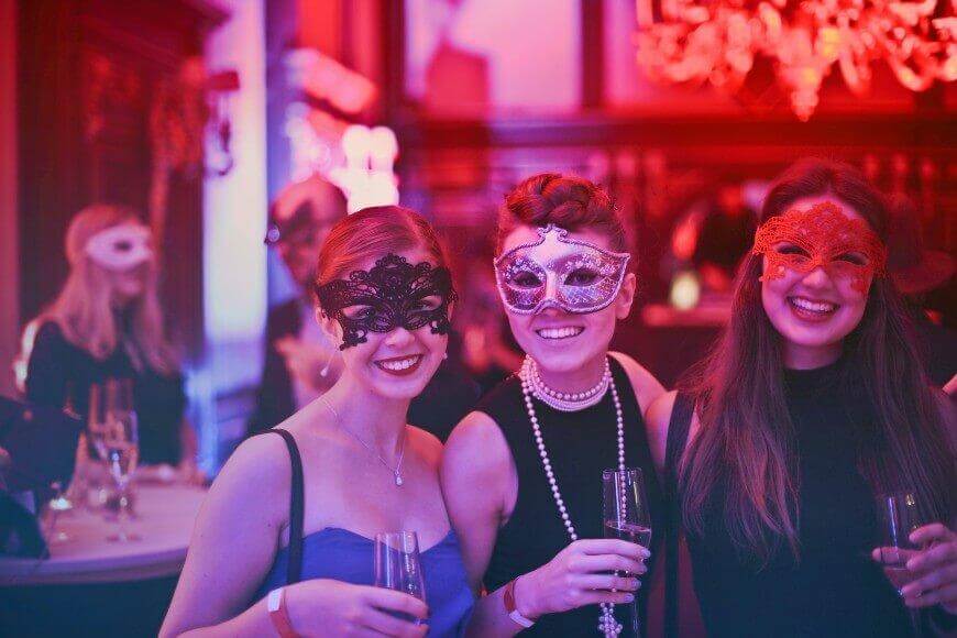 Three women in elegant dresses and decorative masks smile while holding drinks at a masquerade party, with warm red and purple lighting and other masked guests in the background.