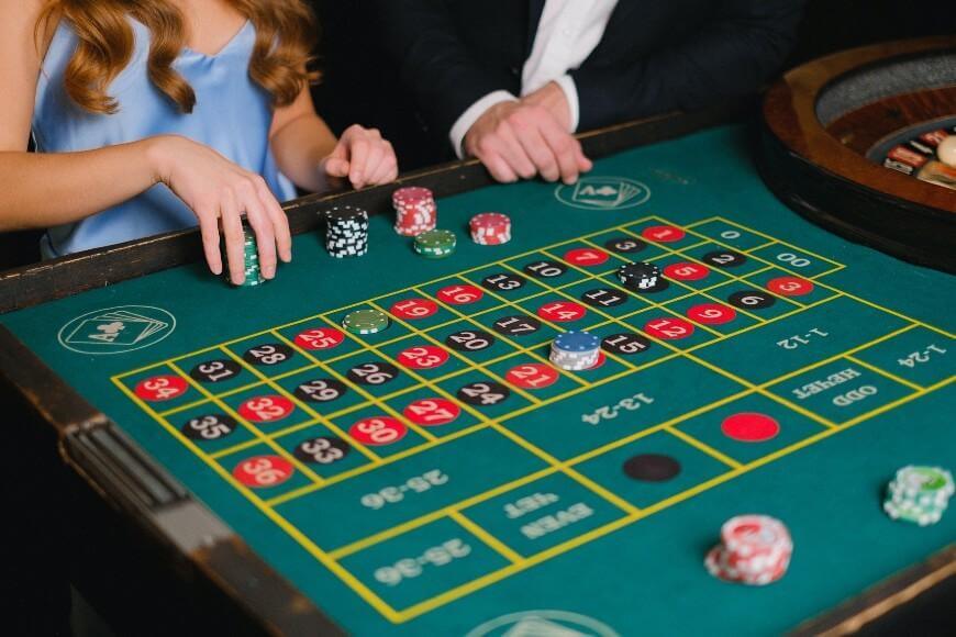 Two people sit at a roulette table, placing chips on numbers. A roulette wheel and stacks of colorful chips are visible, and both are dressed in formal attire.