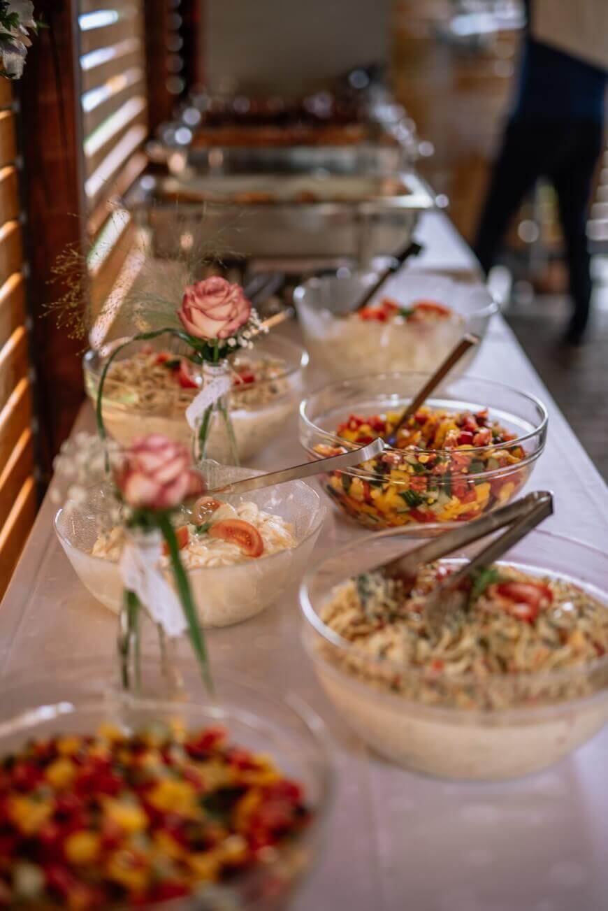 A buffet table with several glass bowls filled with colorful salads and pasta dishes, each with serving utensils. The table is decorated with small vases holding pink roses. Warm light filters in from wooden blinds.