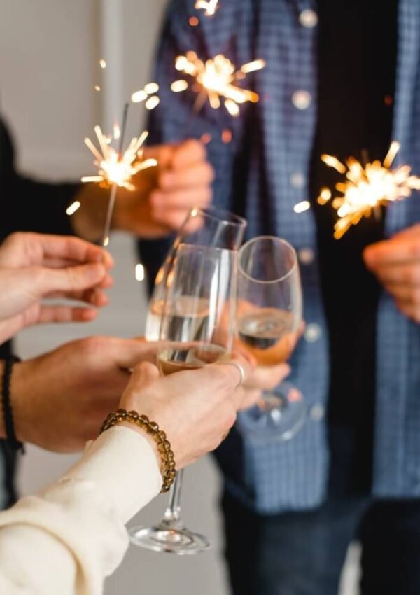 Three people clink glasses of sparkling wine and hold lit sparklers, celebrating together. The focus is on their hands, with festive, warm lighting in the scene.