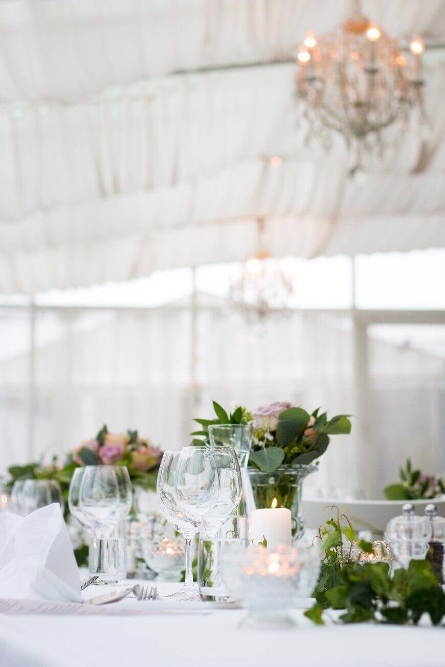 Elegant table setting with empty wine glasses, floral centerpieces, and a lit candle on a white tablecloth. Chandeliers hang overhead, and sheer drapes create a soft, romantic atmosphere.