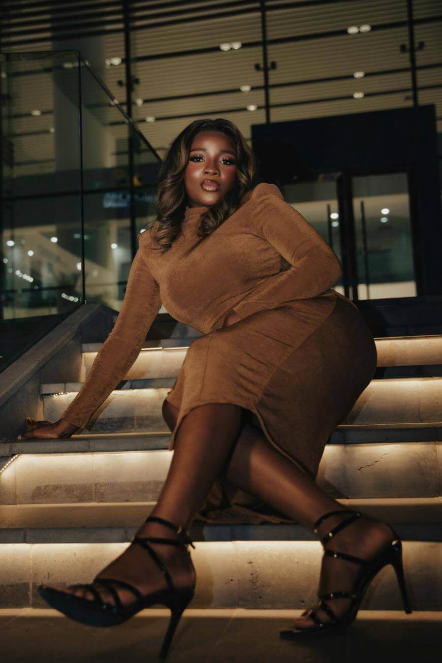 A woman in a fitted brown dress and black strappy heels sits confidently on illuminated stairs at night, with a modern building in the background. She has styled hair and a poised, elegant expression.