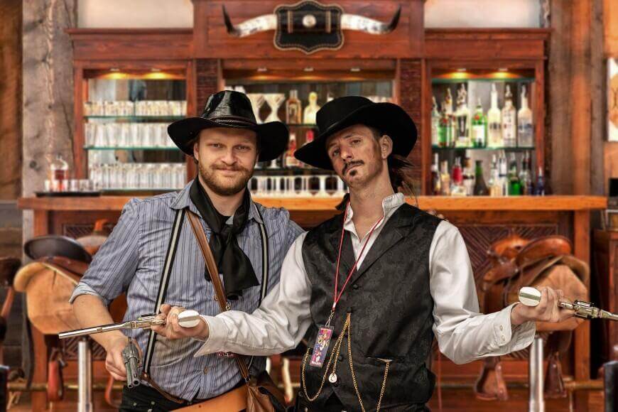 Two men dressed in cowboy costumes pose in front of a bar. Both wear black hats; one has fake guns drawn while the other smiles with his arm around him. The bar and saddle stools are visible in the background.