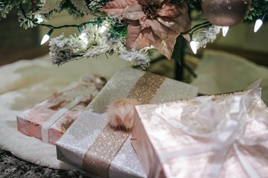 Three glittery, pastel-colored wrapped gifts sit under a Christmas tree decorated with white lights and pink poinsettias. The presents are adorned with shiny ribbons, bows, and a fuzzy pom-pom.