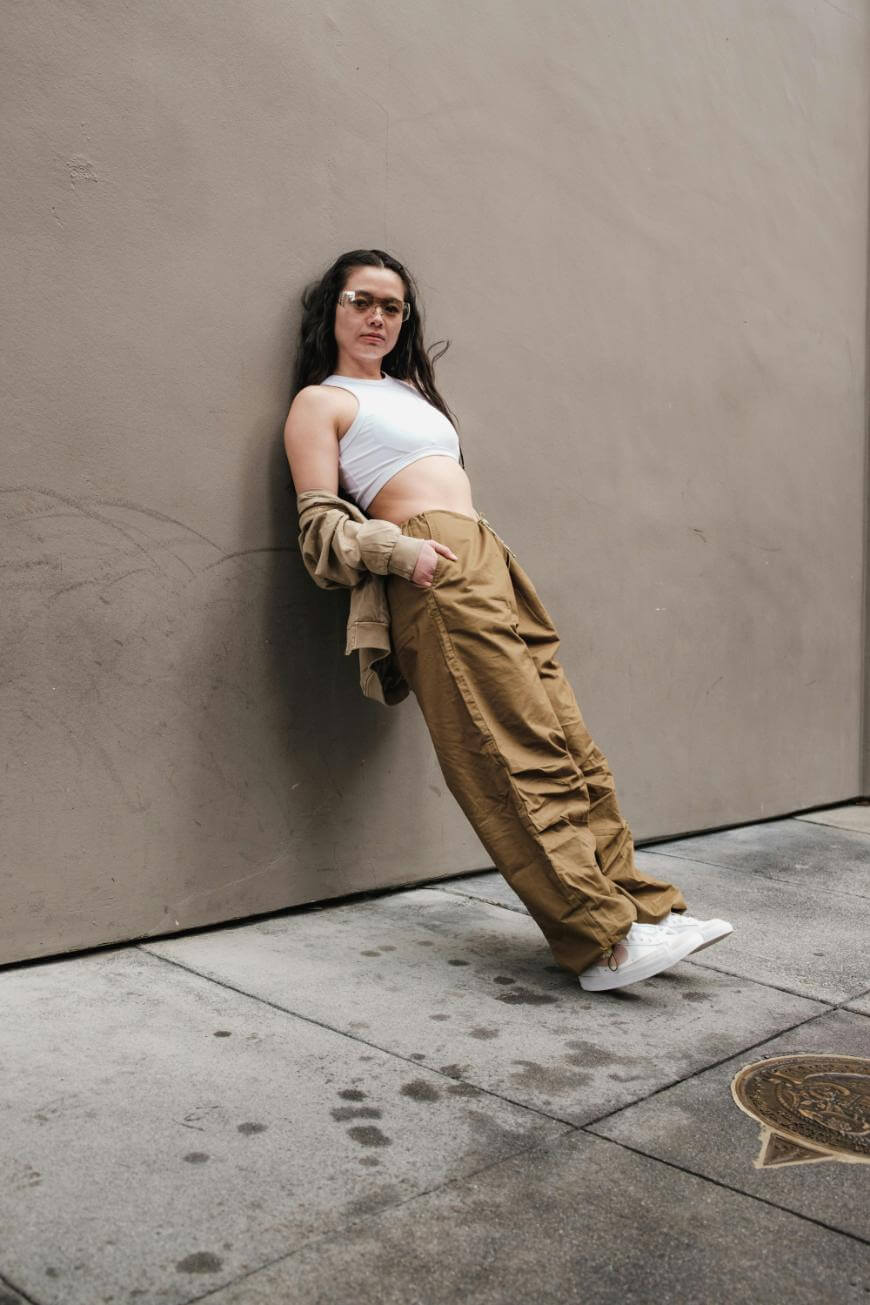 A woman in a white crop top, tan cargo pants, and white sneakers leans back against a beige wall on a city sidewalk, looking confidently at the camera.
