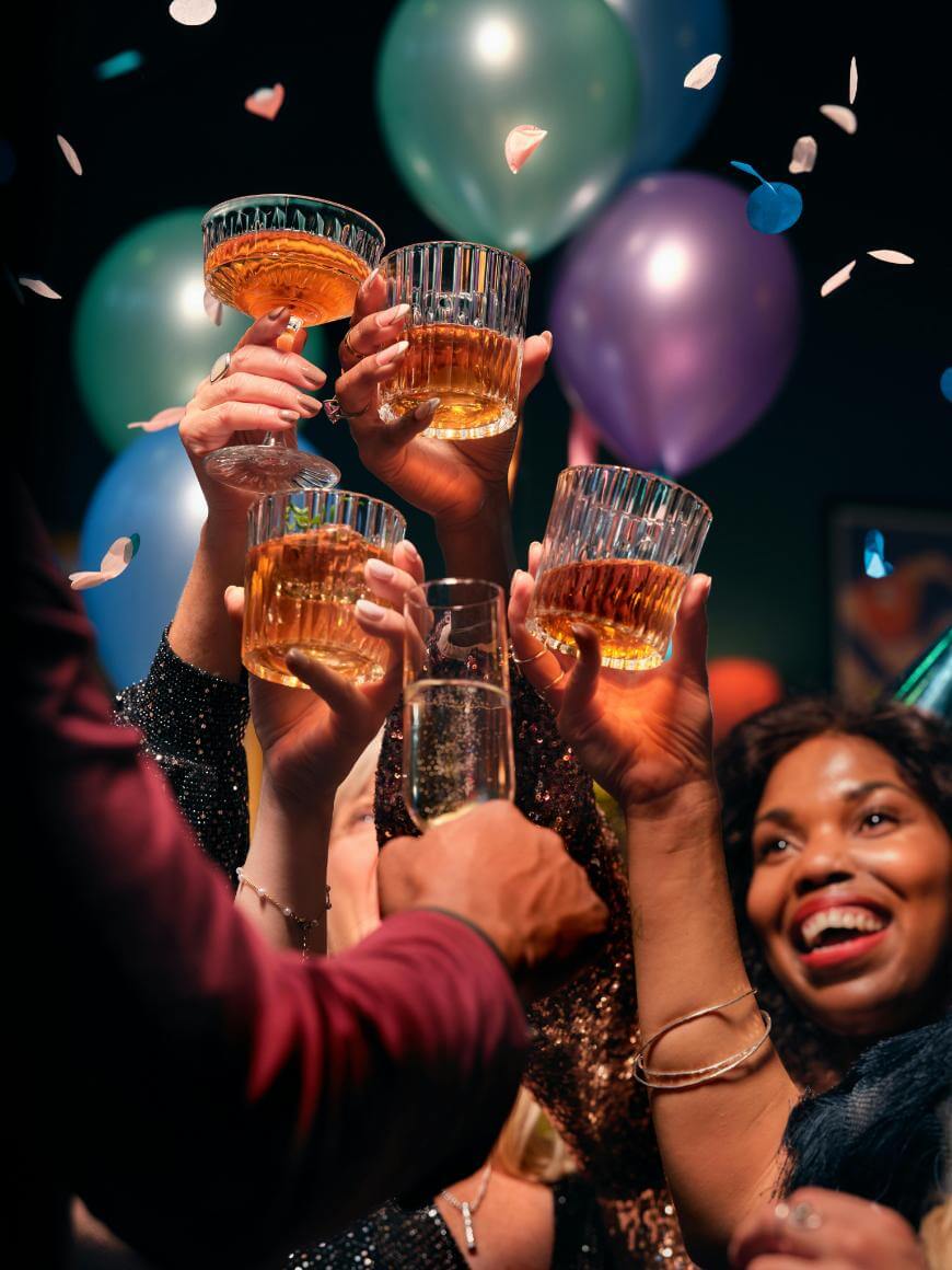 A group of people raise glasses of whiskey and champagne in a celebratory toast, surrounded by colorful balloons and falling confetti, with one woman smiling brightly in the foreground.