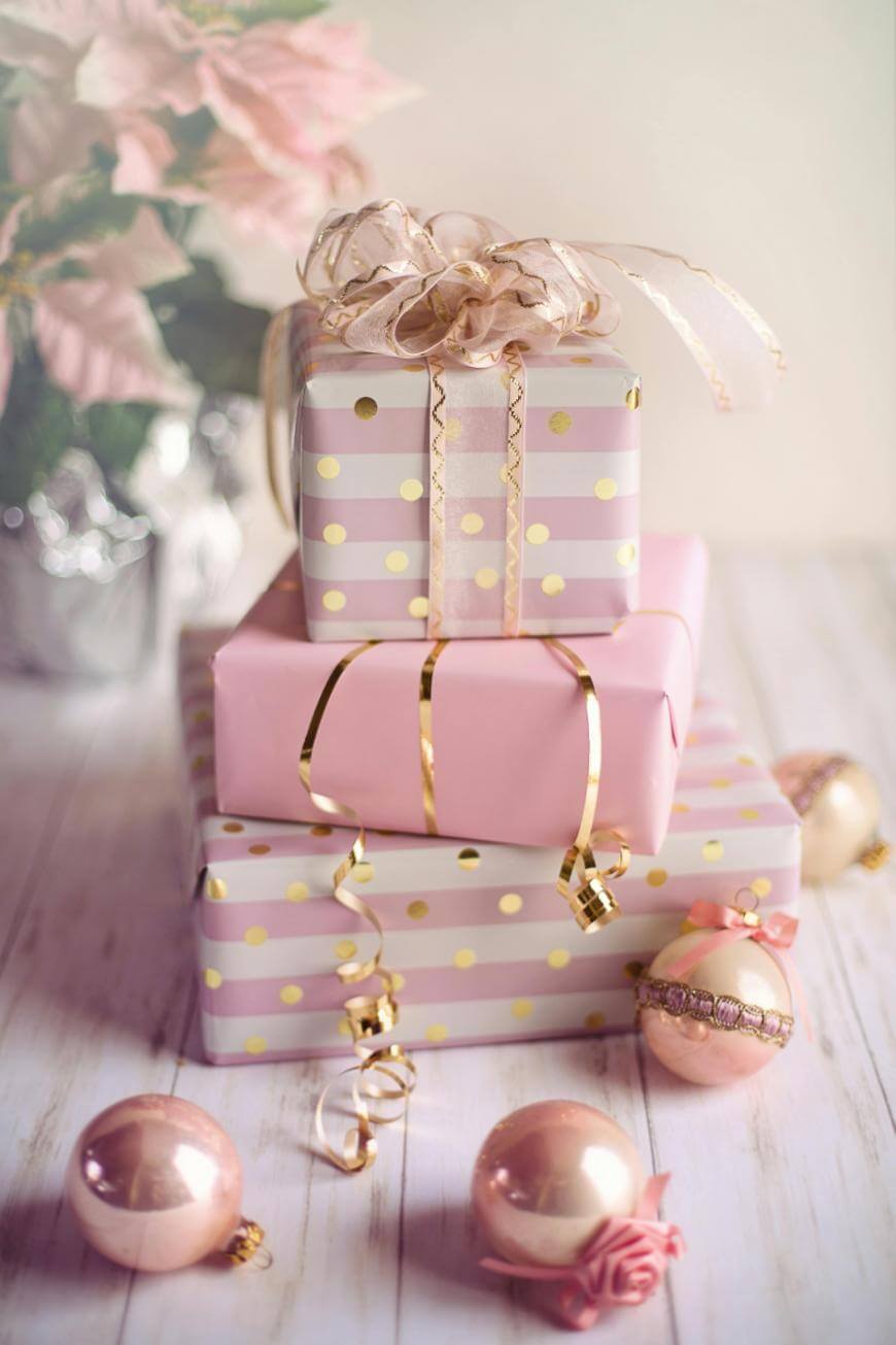 Three pastel pink and gold-wrapped gifts, topped with a ribbon, sit on a white wooden surface, surrounded by gold and pink ornaments and a vase of pink flowers in the background.