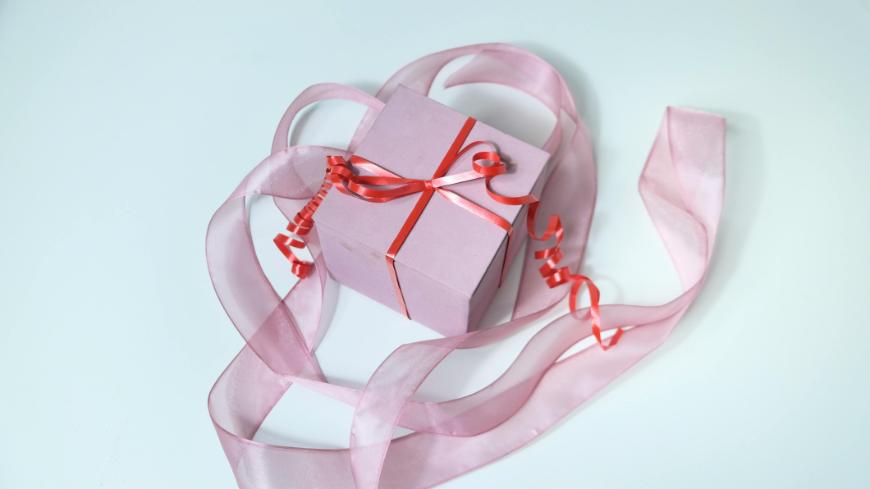 A pink gift box tied with a red ribbon sits on a white surface, surrounded by a long, sheer pink ribbon arranged in a loose heart shape.